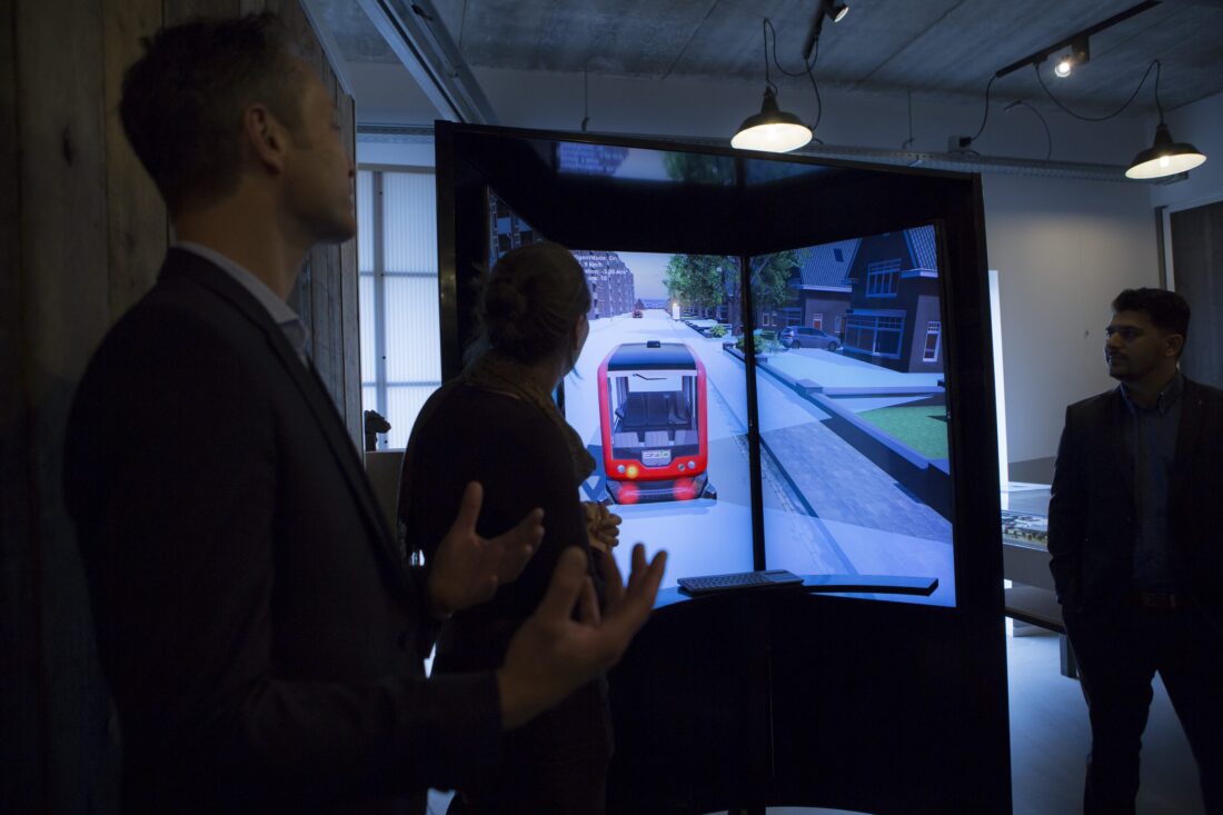 Een groep mensen staat voor een simulator waarop een animatie van een zelfrijdend busje die door de straat rijdt is te zien.