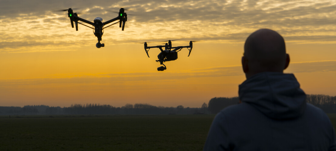 Een man in een grijze hoodie kijkt naar twee drones in de lucht.