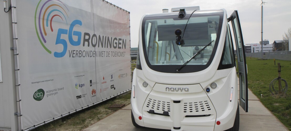 Een witte zelfrijdende shuttlebus van het Franse bedrijf NAVYA geparkeerd naast een banner van 5Groningen: verbonden met de toekomst.
