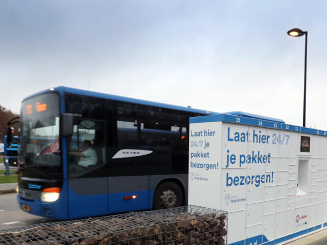 Op de voorgrond kluisjes voor het halen en bezorgen van pakketjes, op de achtergrond rijd een Qliner bus.