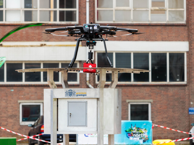 Een closeup van een drone, met daaronder een eerste hulp kit, op een platform. Op de achtergrond een gebouw.