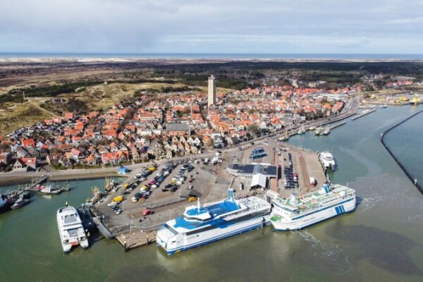 Luchtfoto van een kustplaats met een haven met twee grote aangemeerde schepen, omliggende gebouwen en smalle straatjes. De stad wordt begrensd door water en groen.