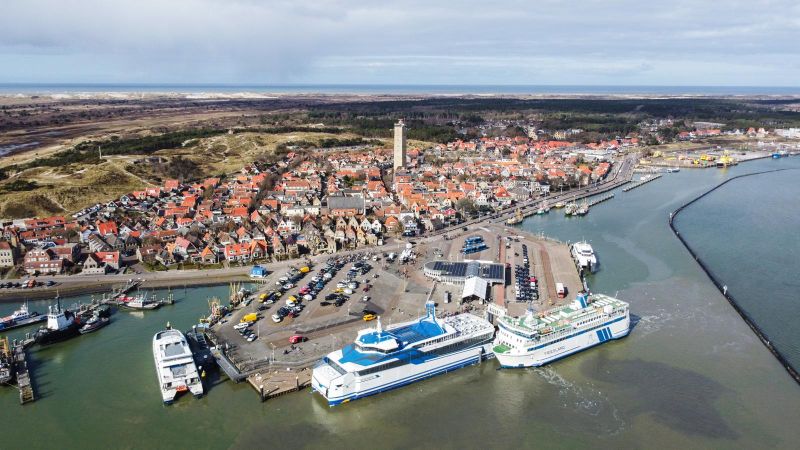 Luchtfoto van een kustplaats met een haven met twee grote aangemeerde schepen, omliggende gebouwen en smalle straatjes. De stad wordt begrensd door water en groen.