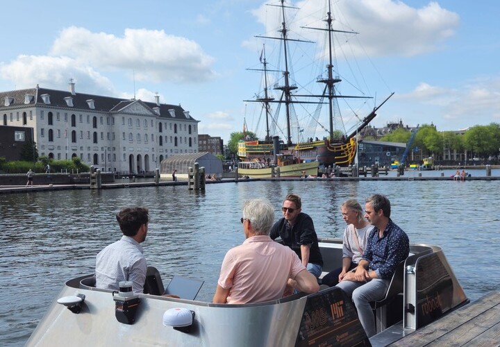 Een groep mensen zit op de Roboat, met op de achtergrond een historisch zeilschip en gebouwen onder een gedeeltelijk bewolkte hemel.