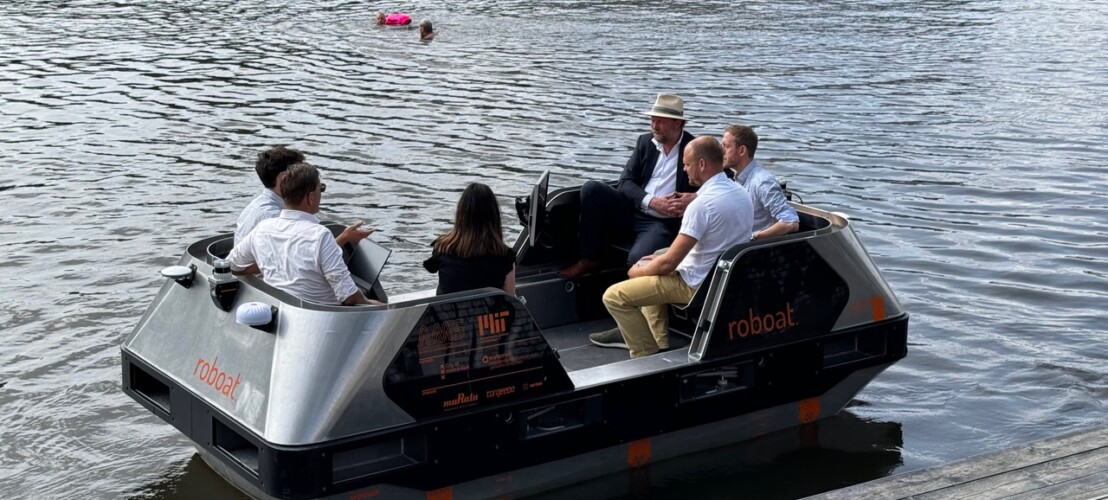 Een groep van zes mensen zit in een klein vaartuig genaamd "Roboat".