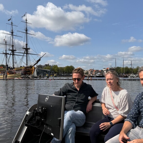 Drie mensen zitten op de roboat met een oud zeilschip op de achtergrond onder een gedeeltelijk bewolkte hemel.