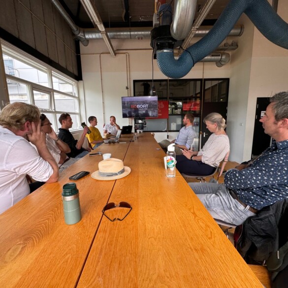 Een groep mensen zit aan een lange houten tafel in een vergaderruimte met grote ramen en een presentatiescherm.