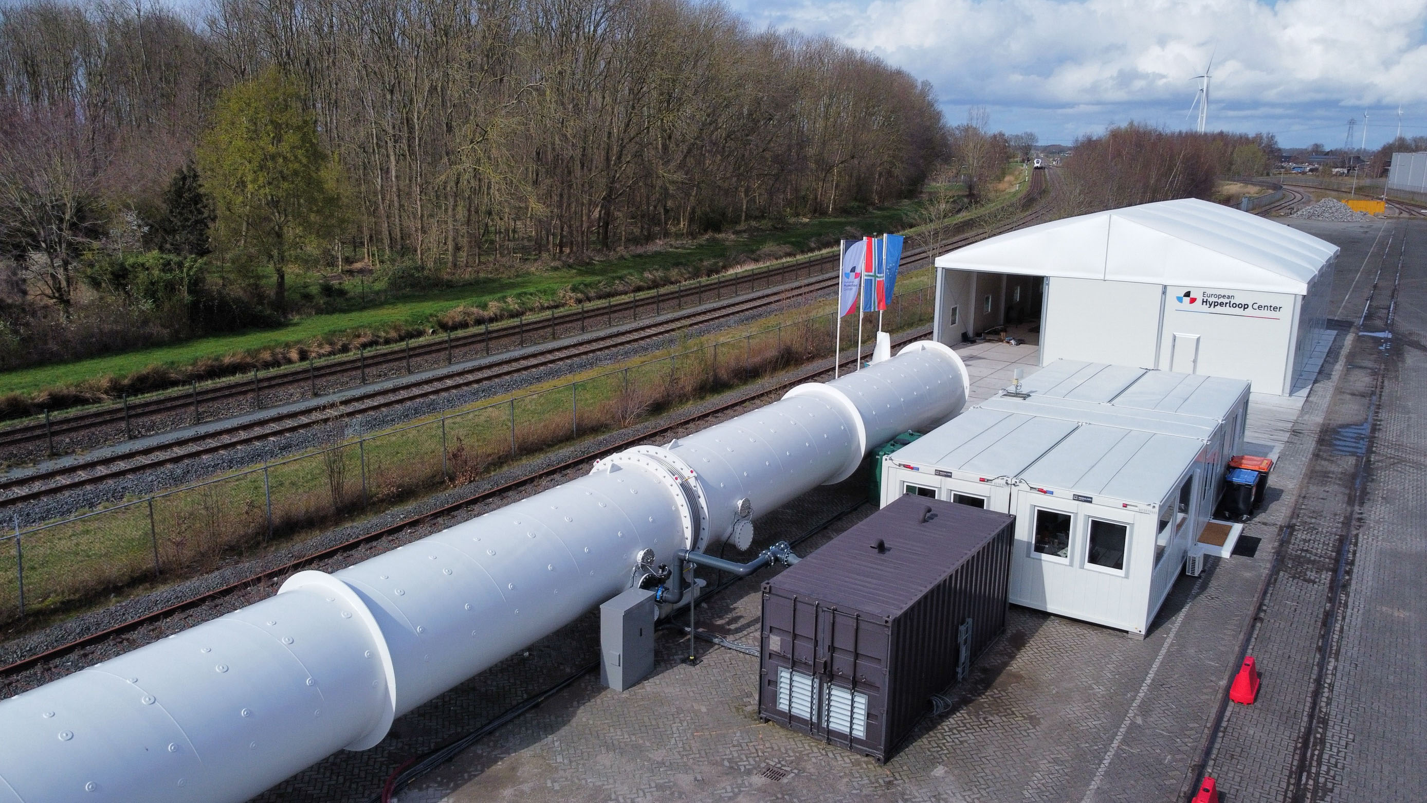 Luchtfoto van een Hyperloop-testfaciliteit met een lange witte buis, containers en nabijgelegen spoorlijnen omgeven door bomen.